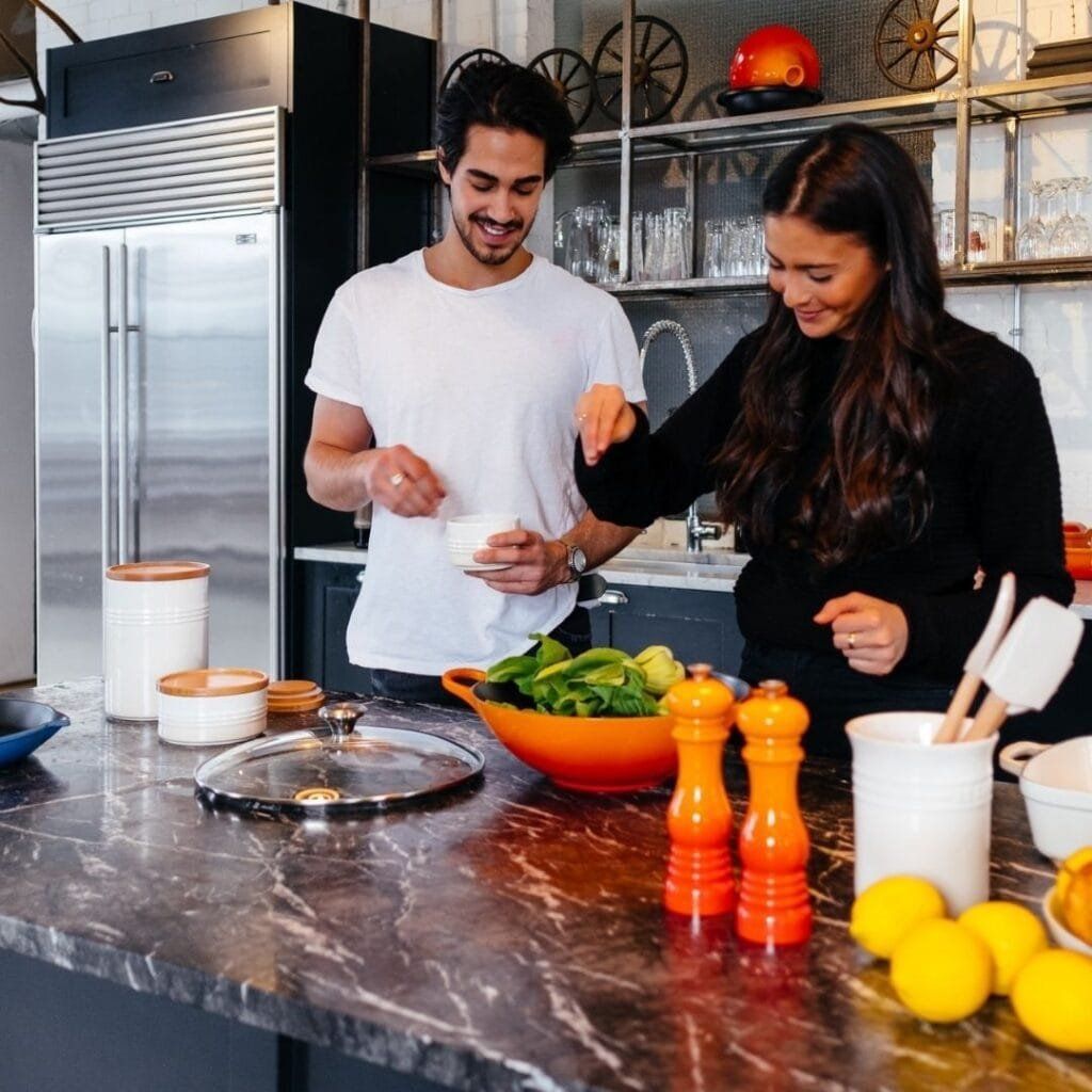 A couple cooking in a modern kitchen. Woman sprinkles seasoning over a salad, man holds a cup.