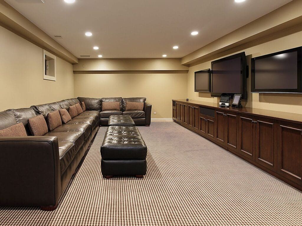 Basement media room with brown leather sectional, built-in media cabinet, three wall-mounted TVs, and patterned carpet.