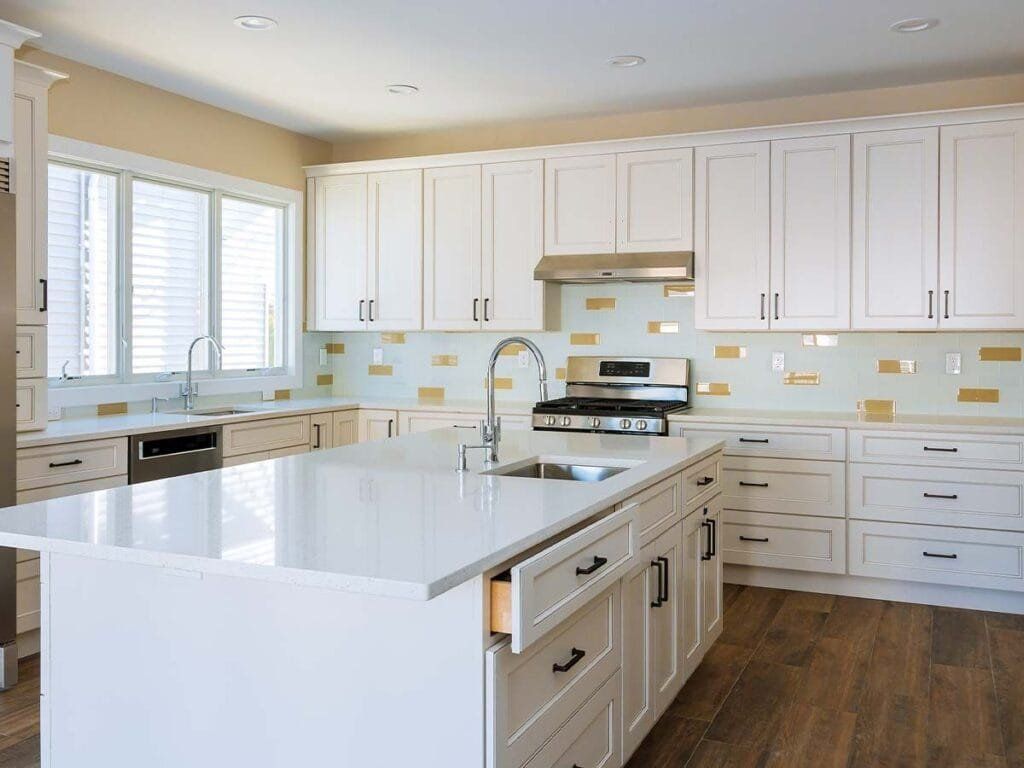 White kitchen with island, cabinets, and appliances. Gold tile backsplash, wooden floor, and a window.