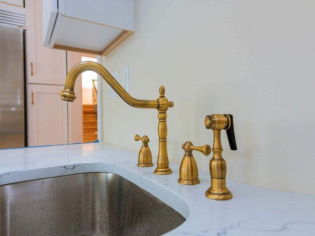 Gold kitchen faucet, sprayer, and controls on a white countertop over a stainless steel sink.