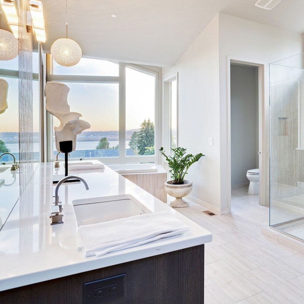 Modern bathroom with white countertops, large window overlooking a body of water, and a glass shower.