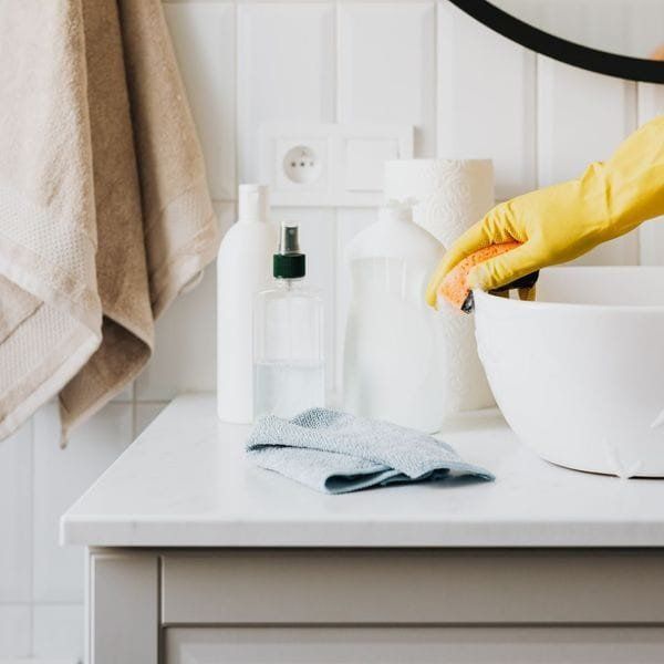 A hand in a yellow glove cleans a white sink. Cleaning supplies and a towel are on the counter.