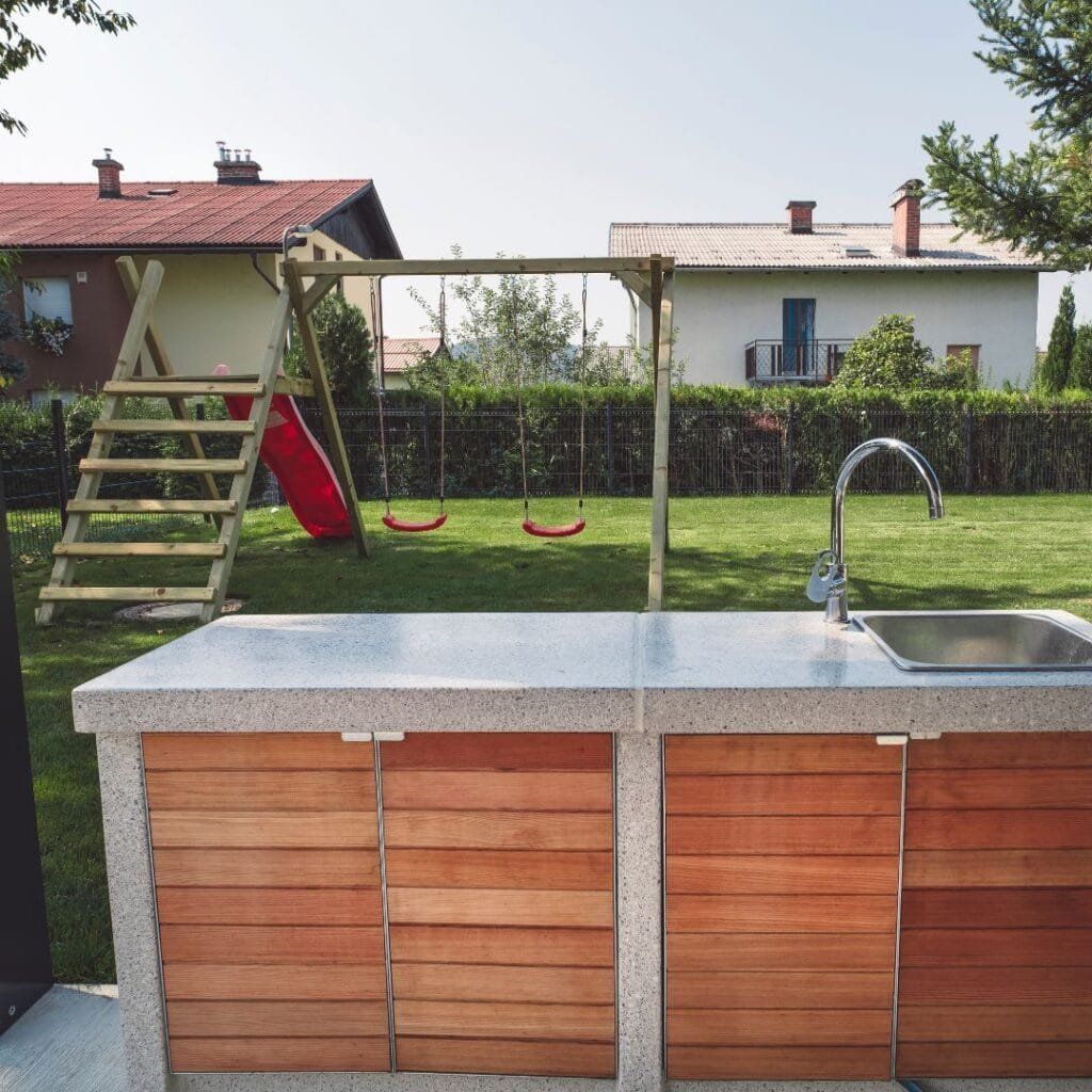 Outdoor kitchen with a wooden facade and speckled countertop, backyard with a swing set and slide.