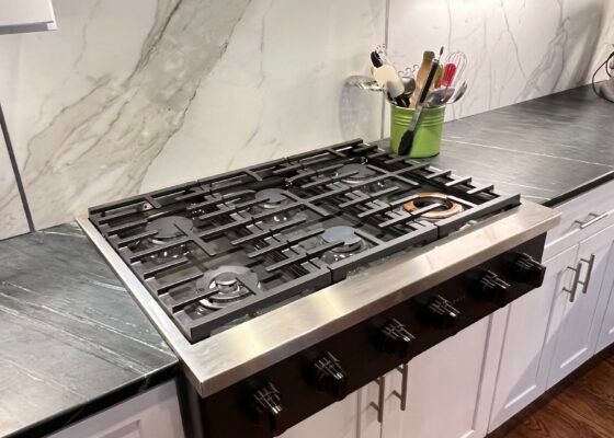 Gas cooktop with six burners, stainless steel, set in black countertop, white cabinets below.