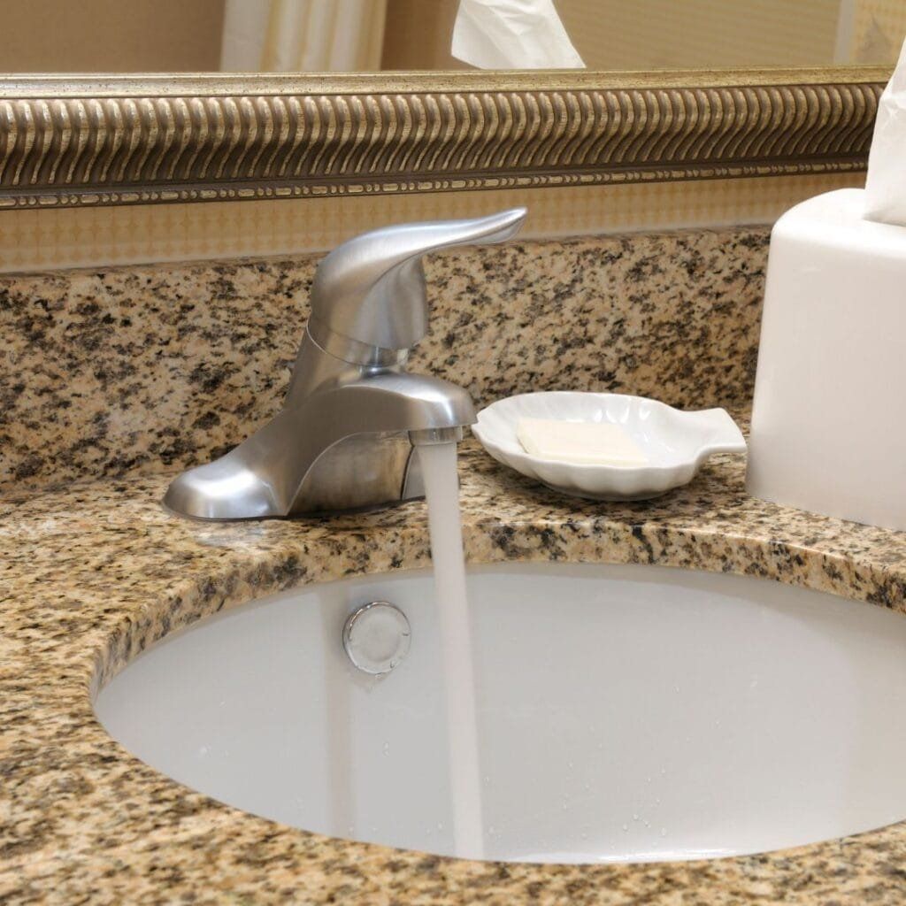 Silver faucet with water running into a white sink on a granite countertop.