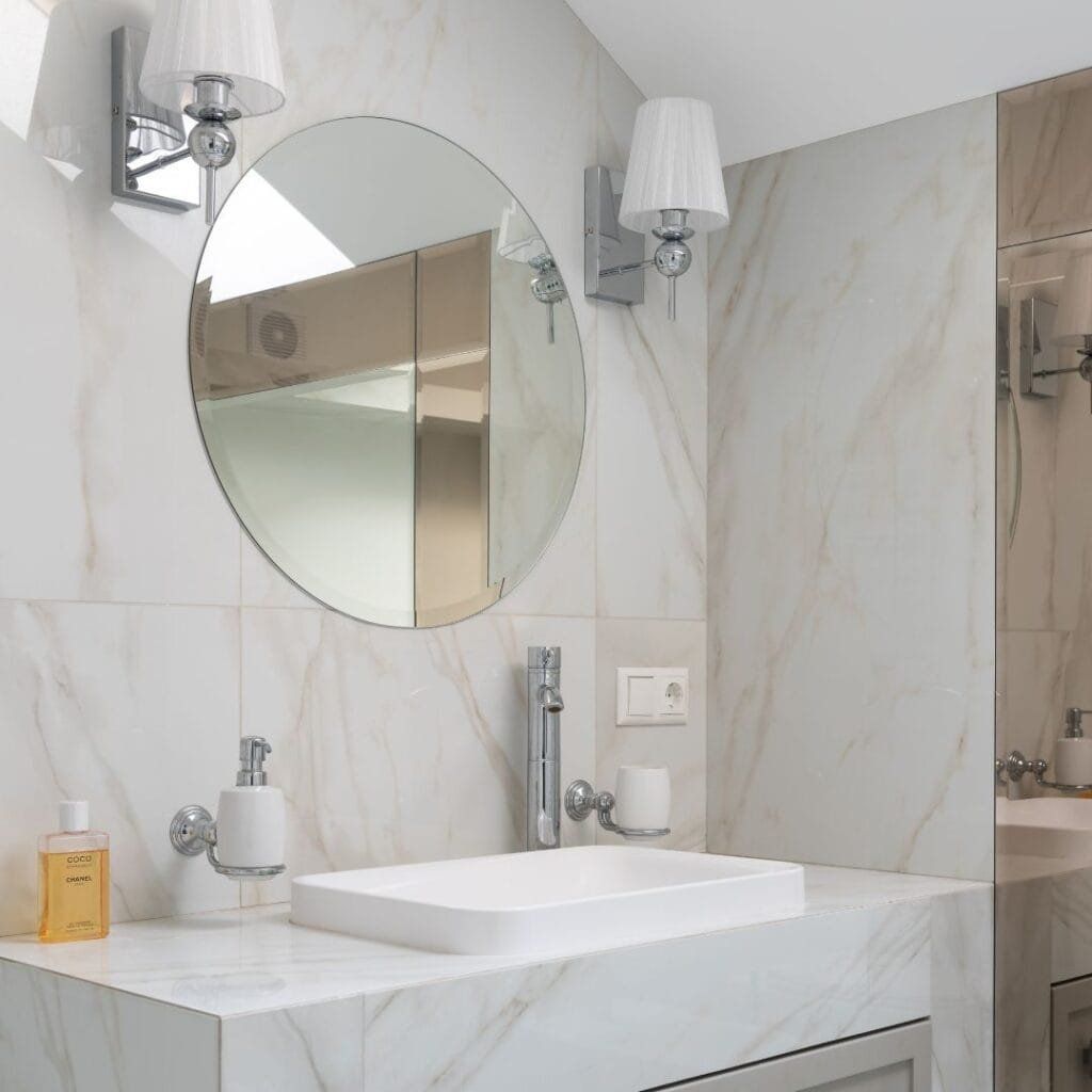 Bathroom with round mirror, white sink, and light-colored marble walls. Two wall sconces flank the mirror.