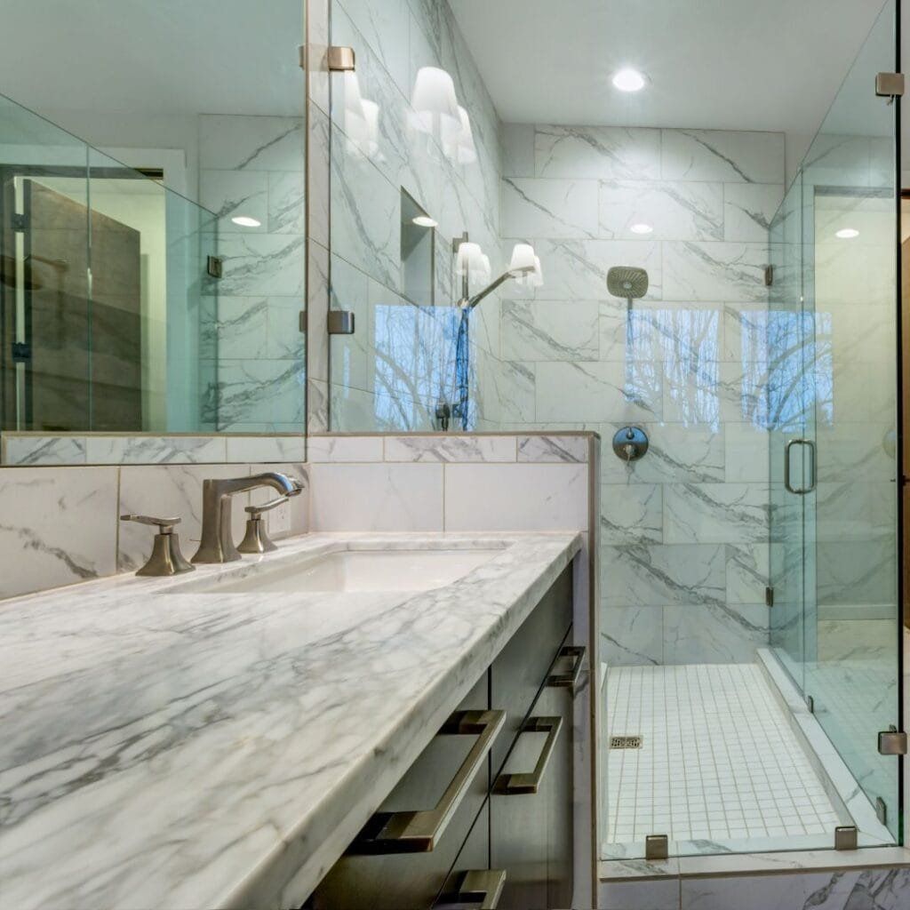 Bathroom with marble-patterned tile and countertop. Glass shower and mirror. Silver faucet and handles.
