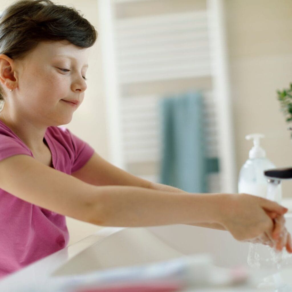 Girl washing hands at a bathroom sink, pink shirt, soap dispenser, and towel rack in background.
