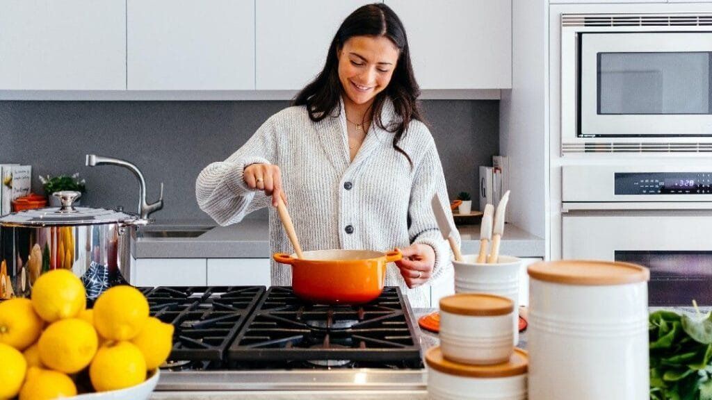 Woman smiles while stirring a pot on a stovetop in a modern kitchen. Lemons, containers, and microwave are visible.