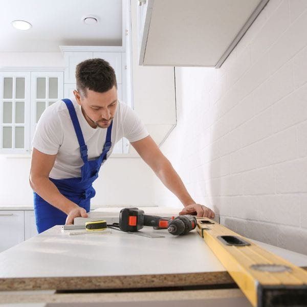 Man in blue overalls using a level and drill on a kitchen countertop.