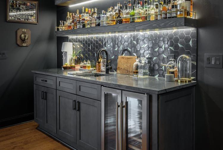 Dark gray home bar with liquor bottles, sink, wine cooler, and hexagon tile backsplash.