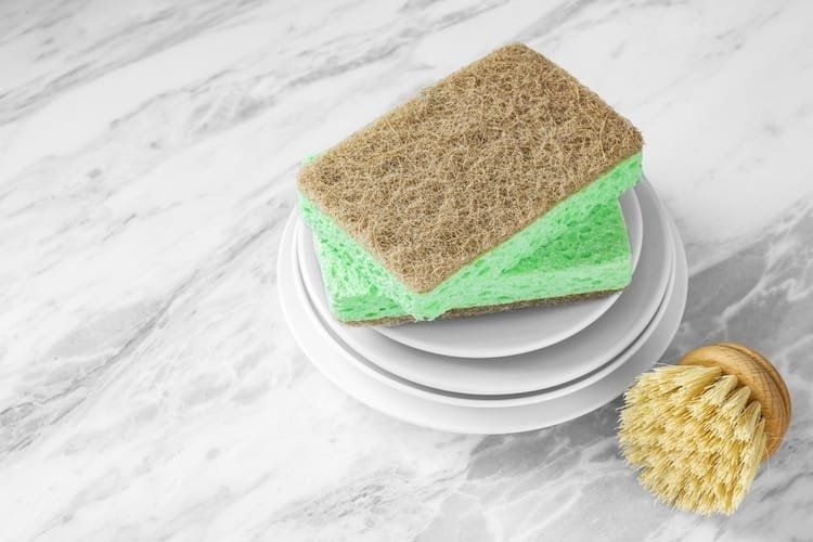 Stack of white plates with green sponges and dish brush on marble surface.