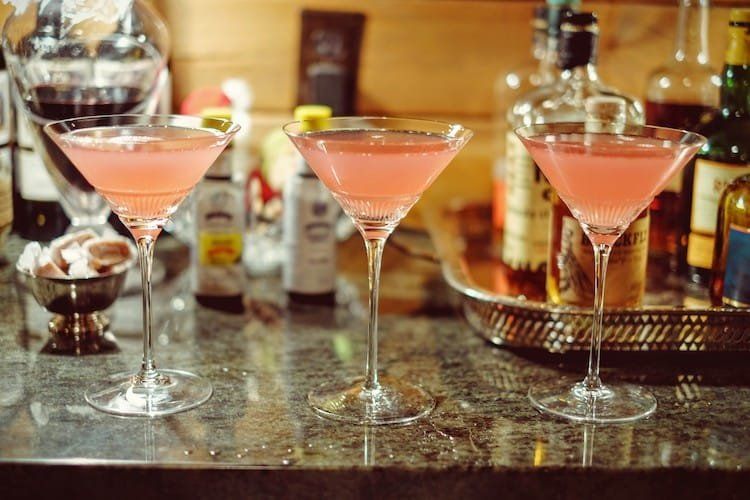 Three pink cocktails in martini glasses on a bar with bottles and glassware in the background.