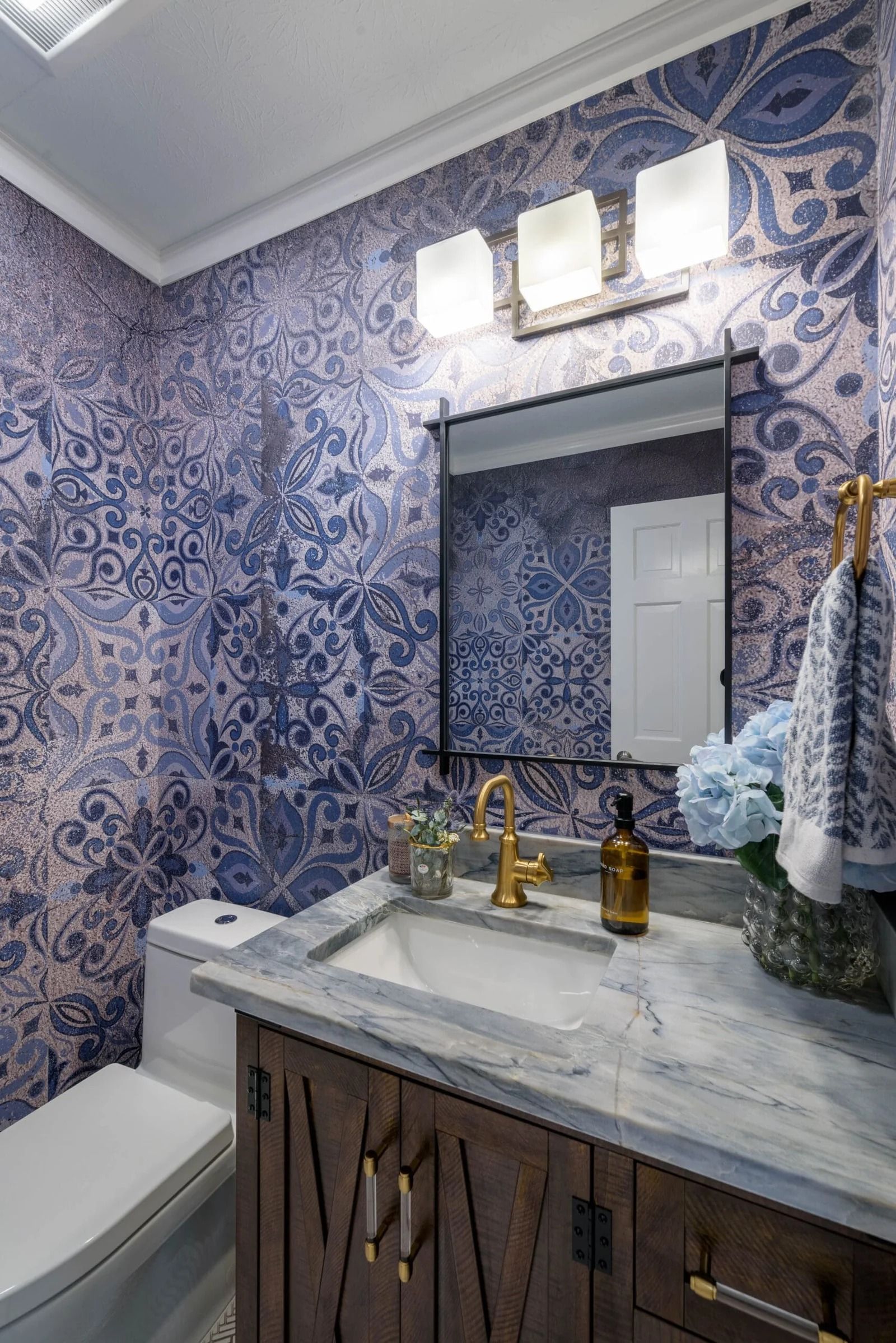 Blue patterned wallpaper bathroom with wooden vanity, gold faucet, and mirror.