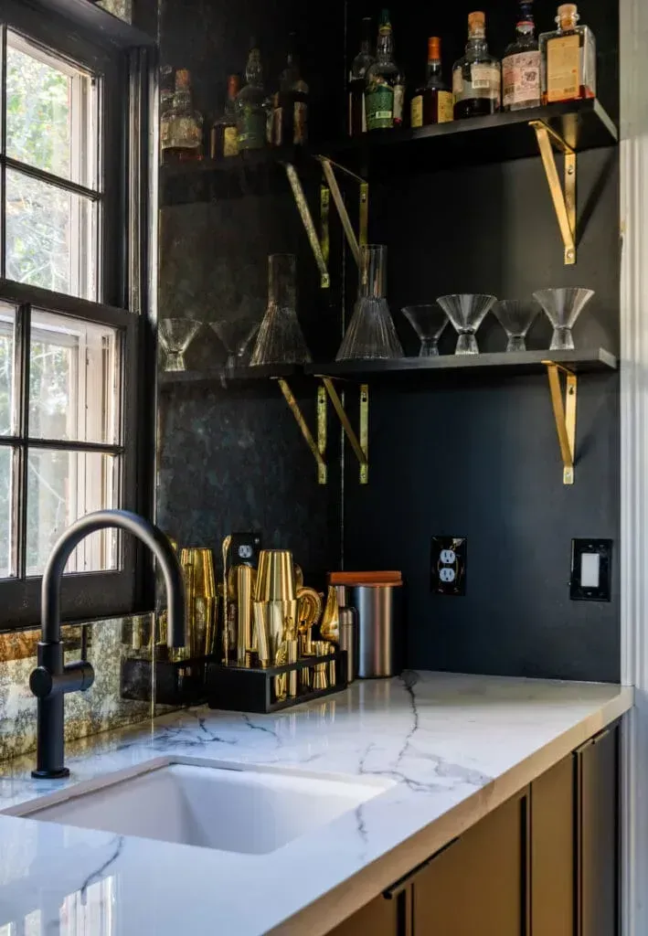 Black and gold home bar with marble countertop, black cabinets, shelves with glassware, bottles, and a sink.
