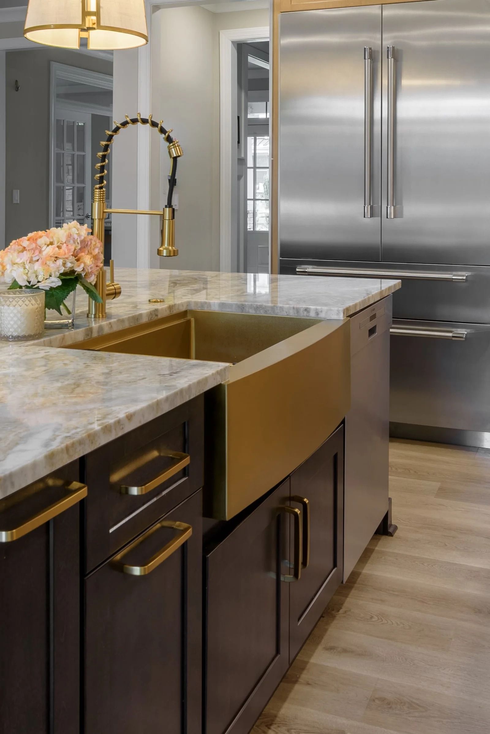 Kitchen island with gold sink, faucet, and hardware, brown cabinets, marble countertop, stainless steel refrigerator.