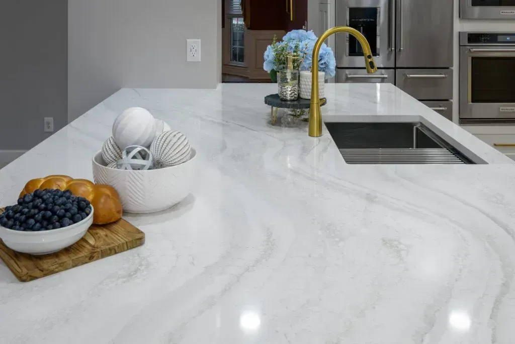 White marble kitchen island with gold faucet, sink, and decorative bowls of fruit and spheres.