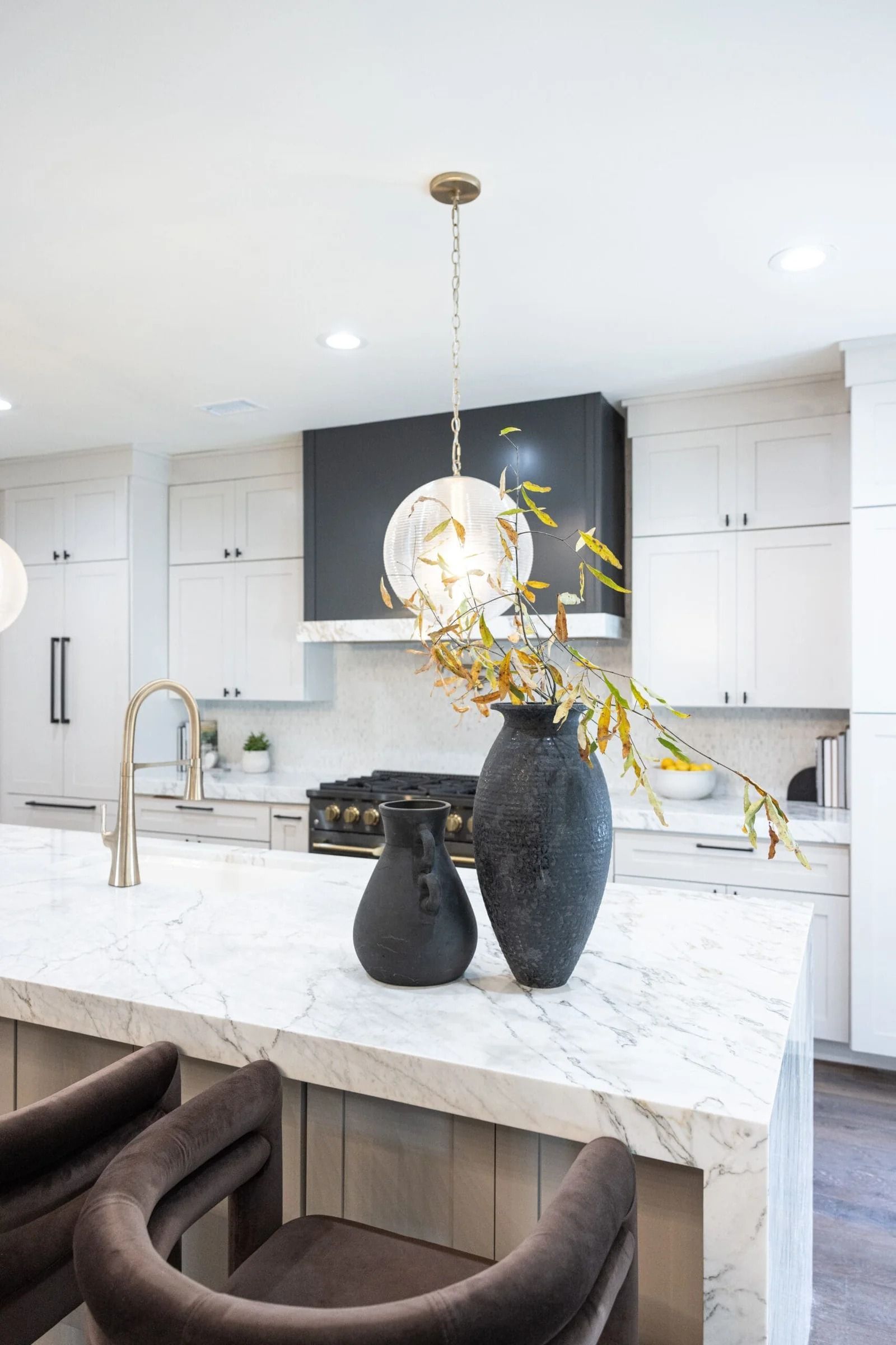 White kitchen with marble island, dark vases, gold light fixture, and bar stools.