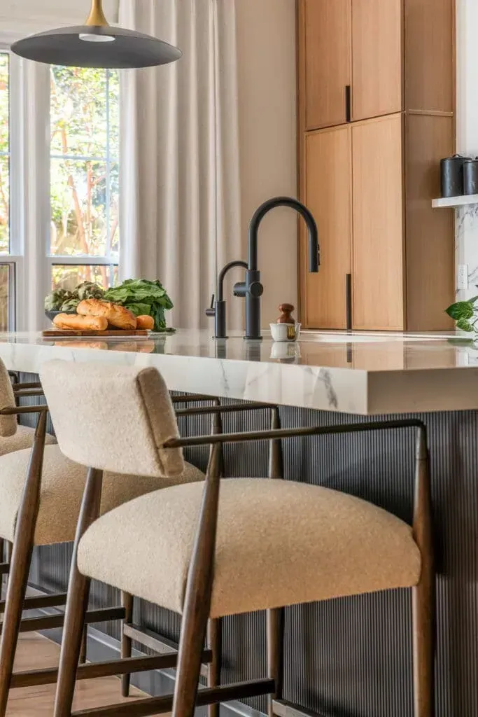 Kitchen island with cream stools, dark faucet, light wood cabinets, and window.