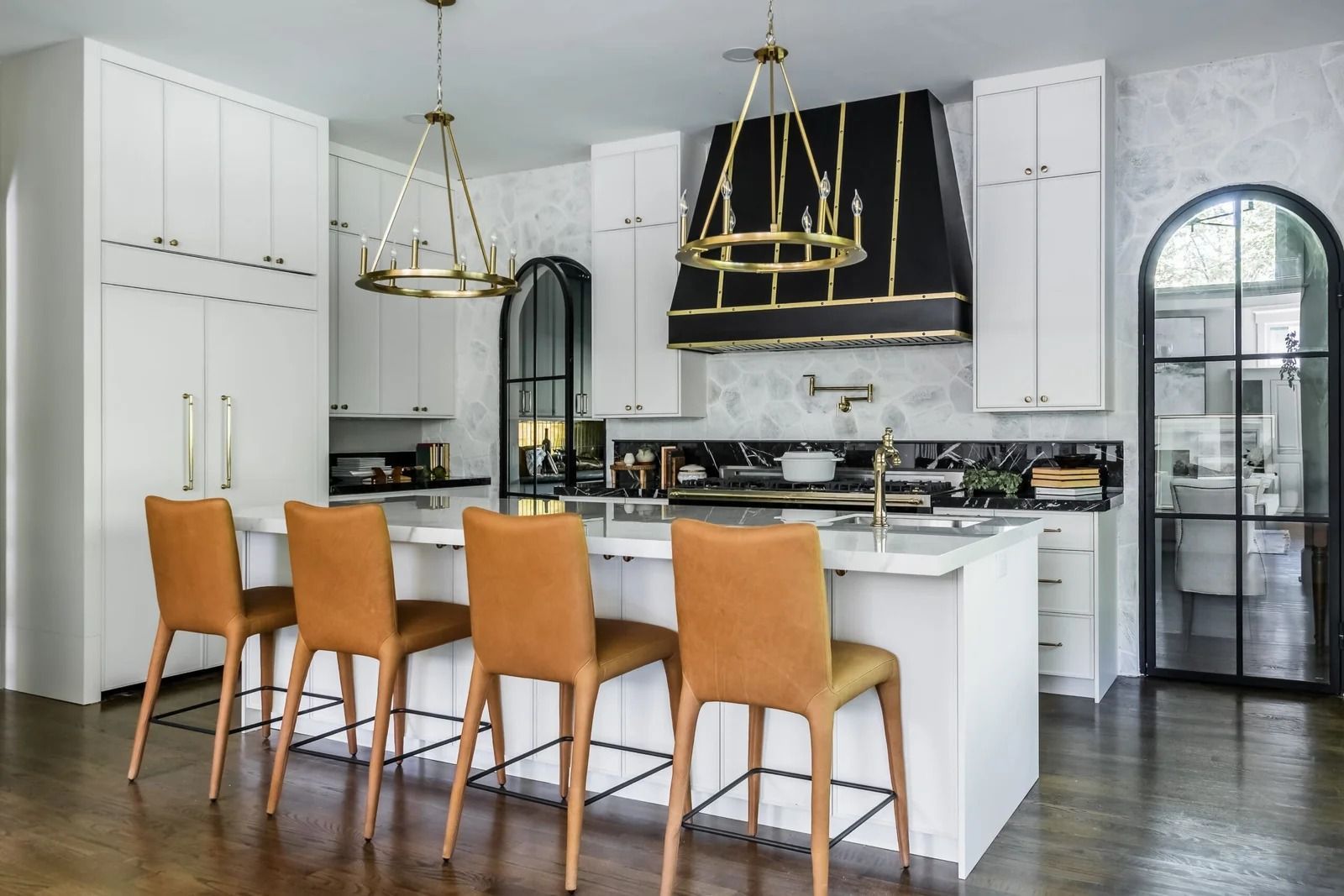 Modern white kitchen with island, brown leather barstools, black range hood, and gold light fixtures.