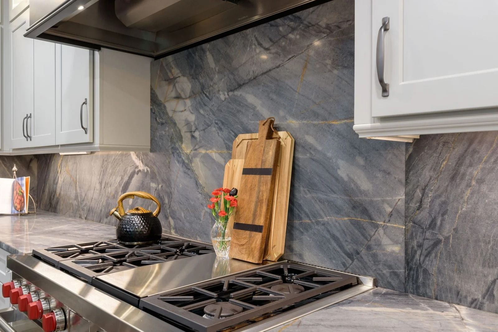 Kitchen with a gray and blue marble backsplash, stovetop, white cabinets, and wooden cutting boards.