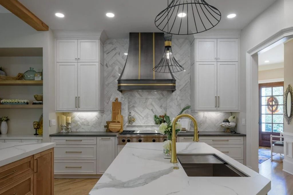 Modern kitchen with white cabinets, marble backsplash, island with gold faucet, and black hood.