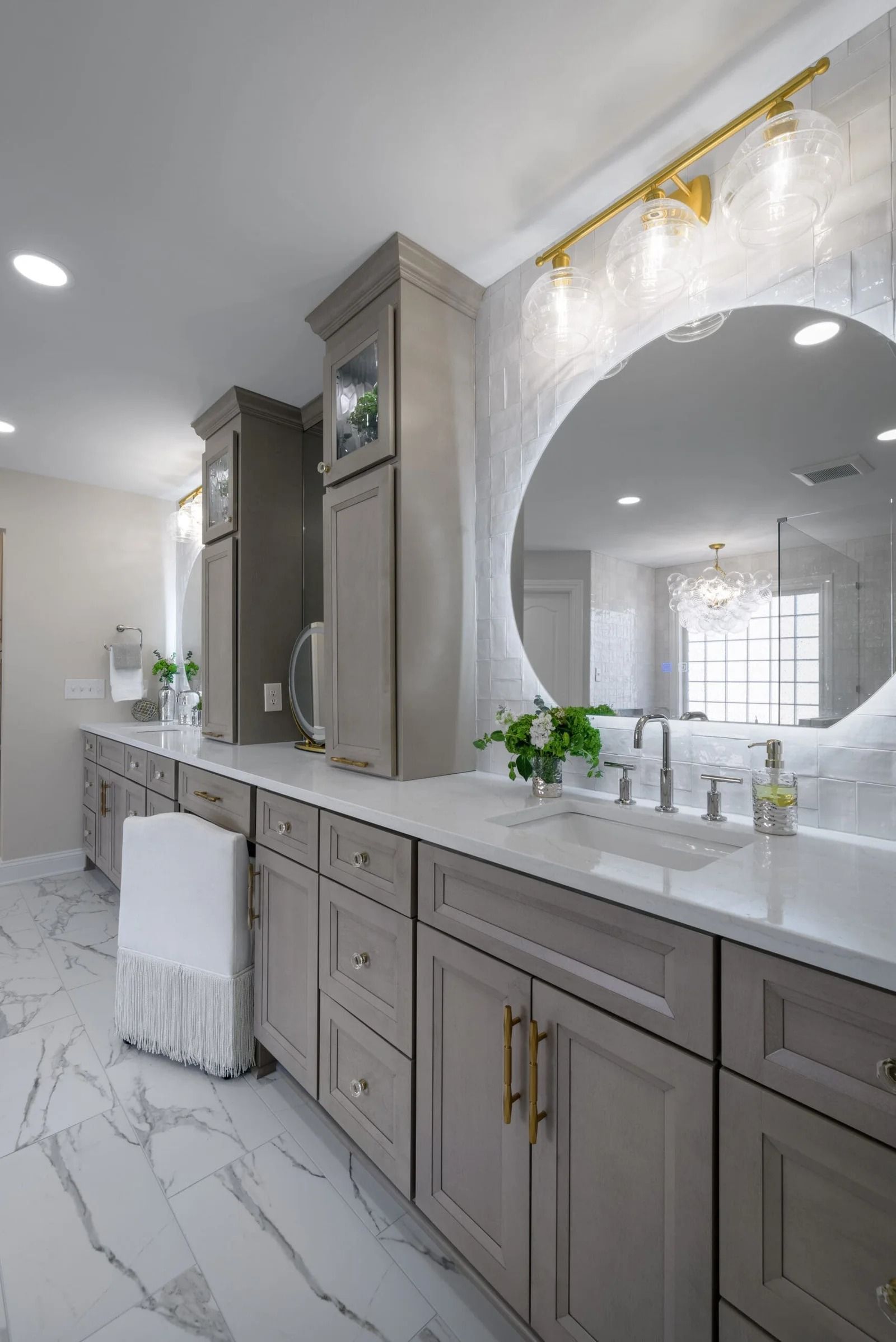 Modern bathroom with gray cabinets, marble floors, and a large round mirror.