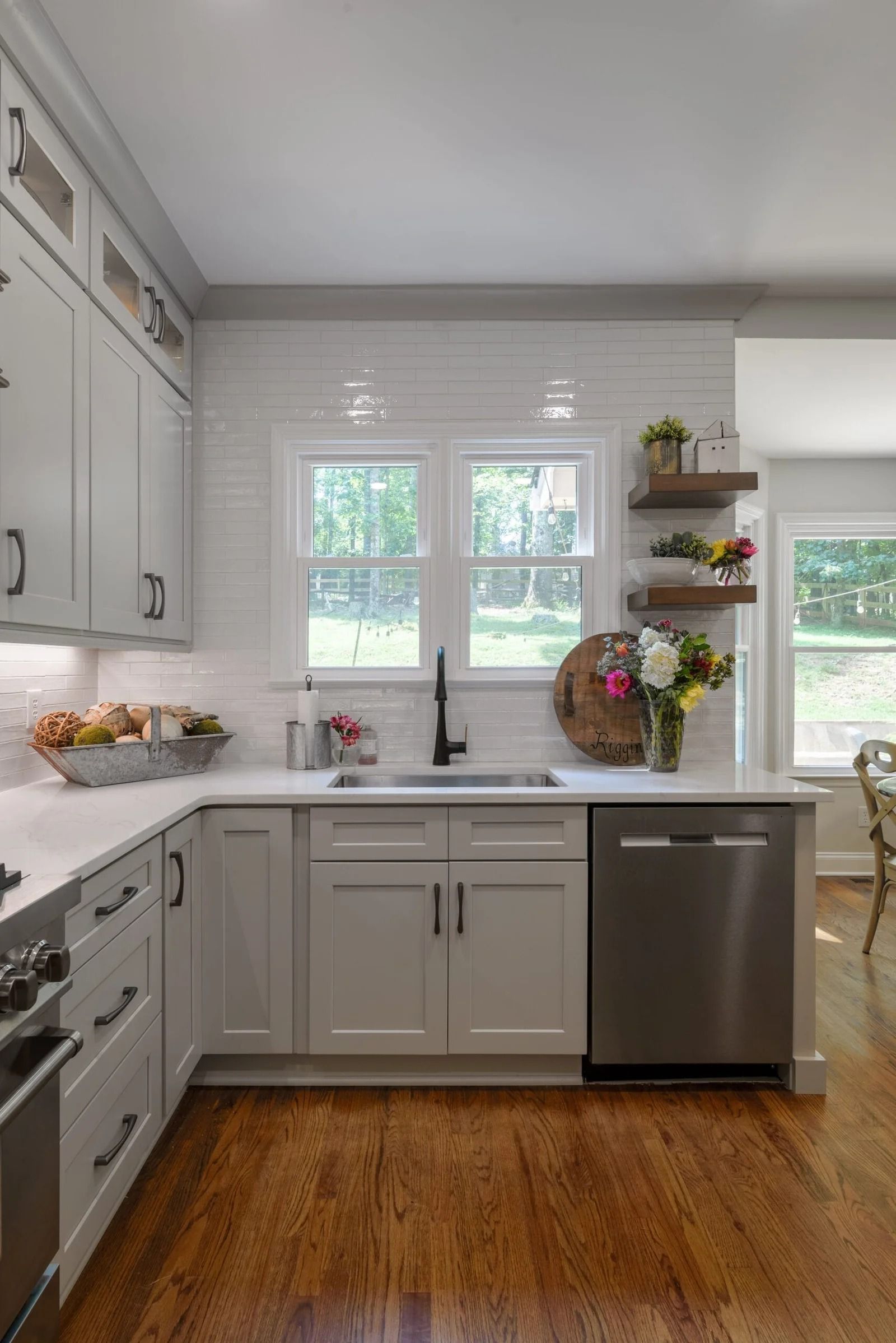 White kitchen with stainless steel appliances, white tile backsplash, and wooden shelves with flowers.