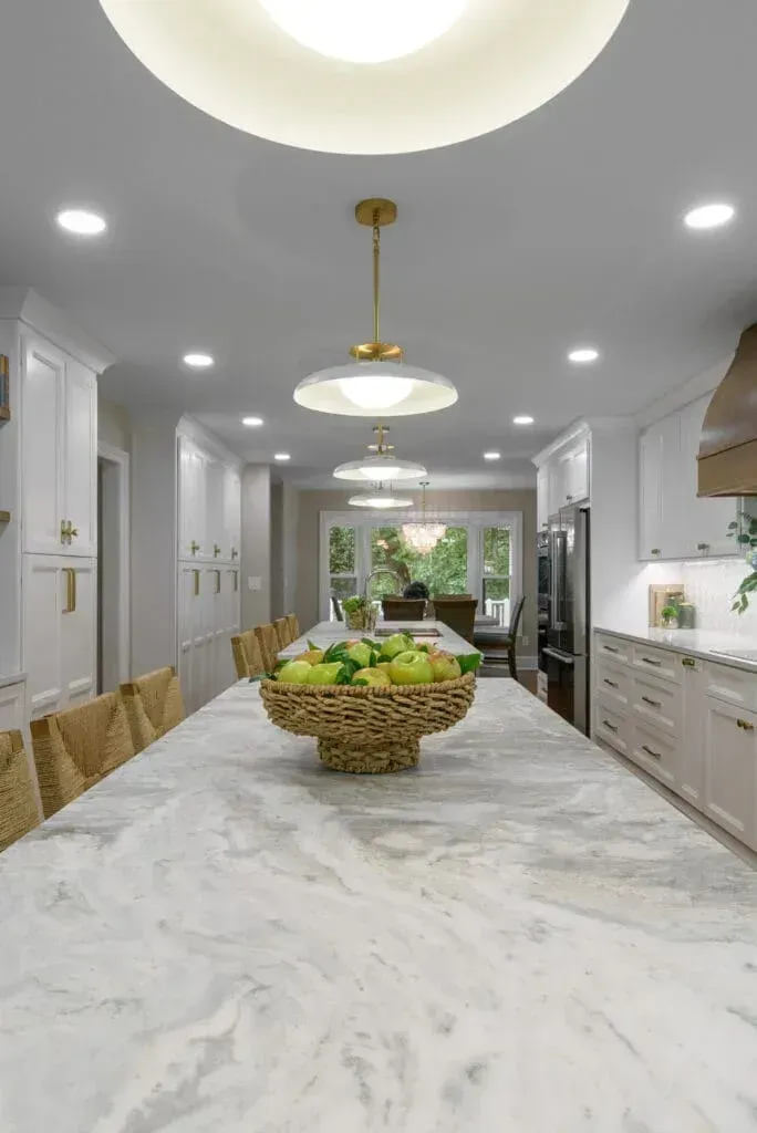 Bright white kitchen with long marble island, fruit basket, pendant lights, and stainless steel appliances.