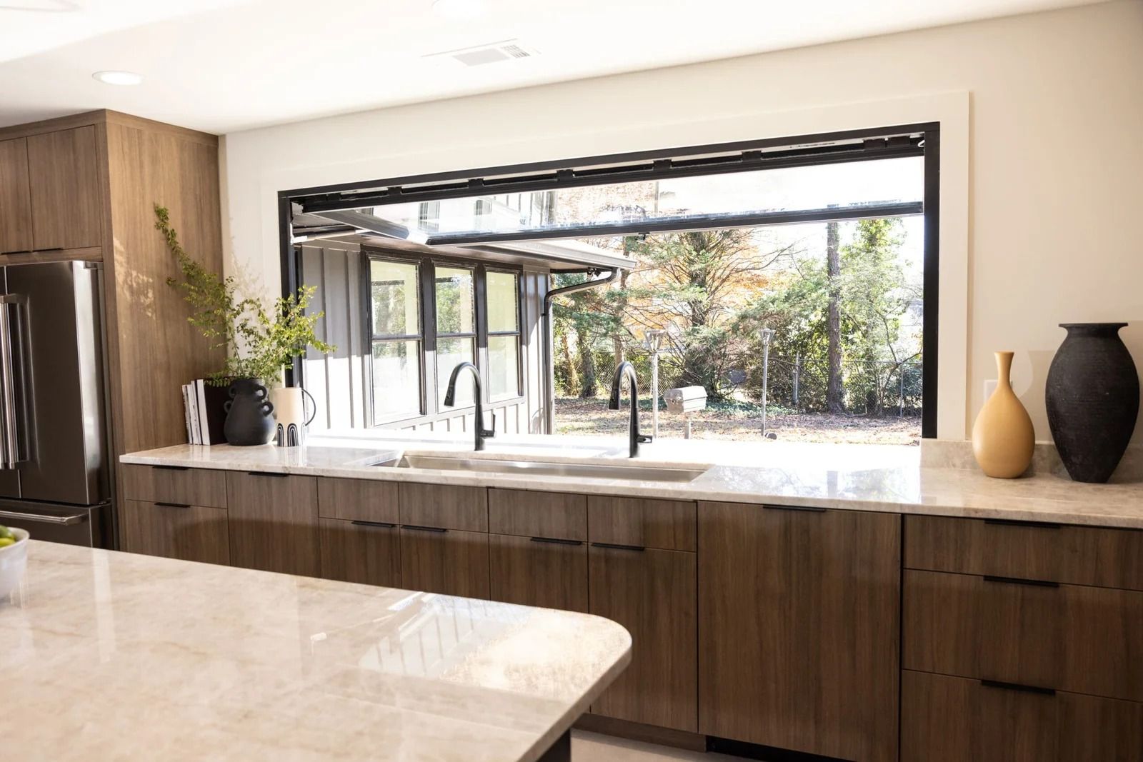 Kitchen with large window overlooking backyard. Brown cabinetry, white countertop, and black fixtures.