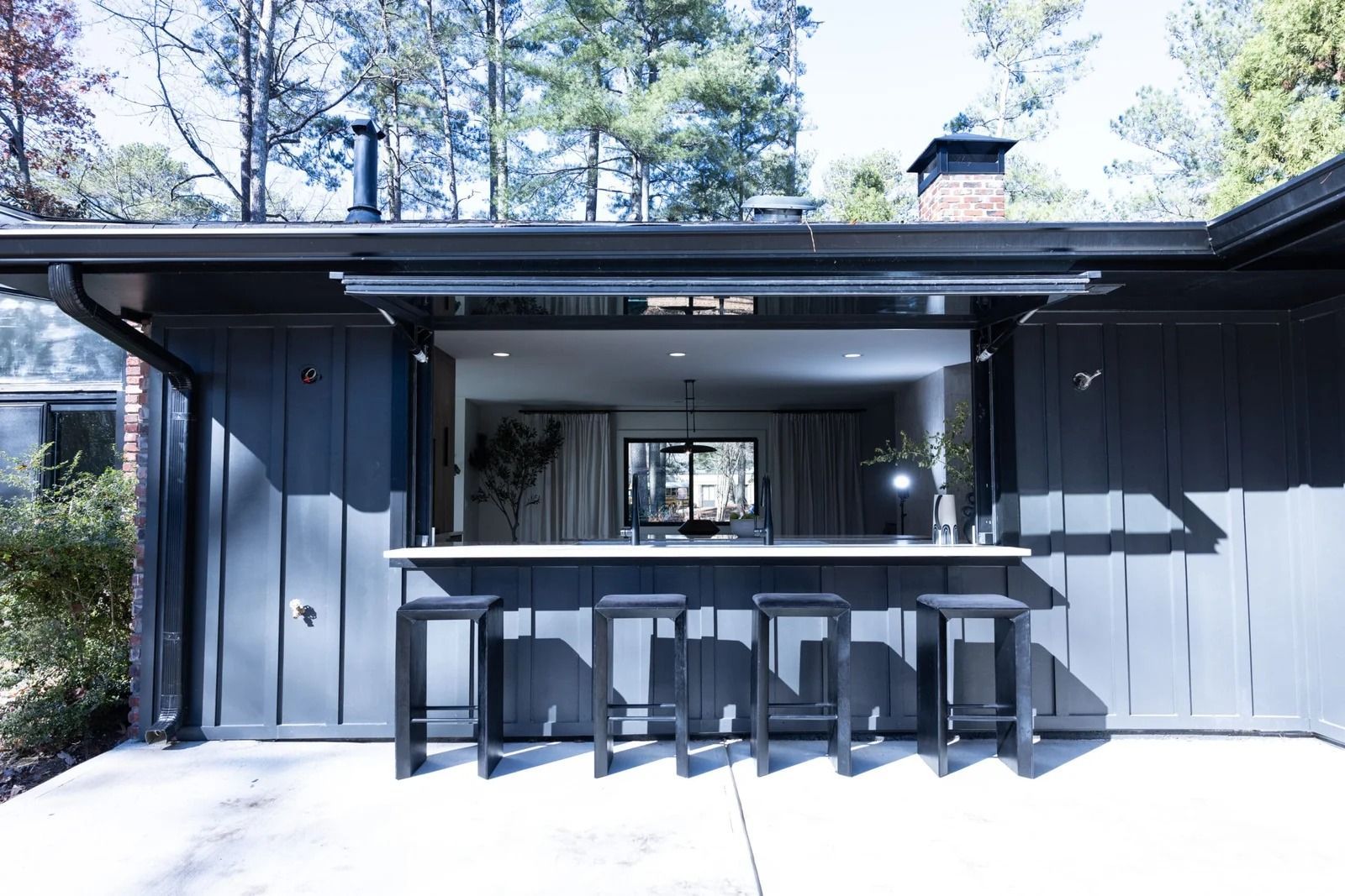 Outdoor bar with barstools; dark gray siding, window opens to interior, trees in the background.