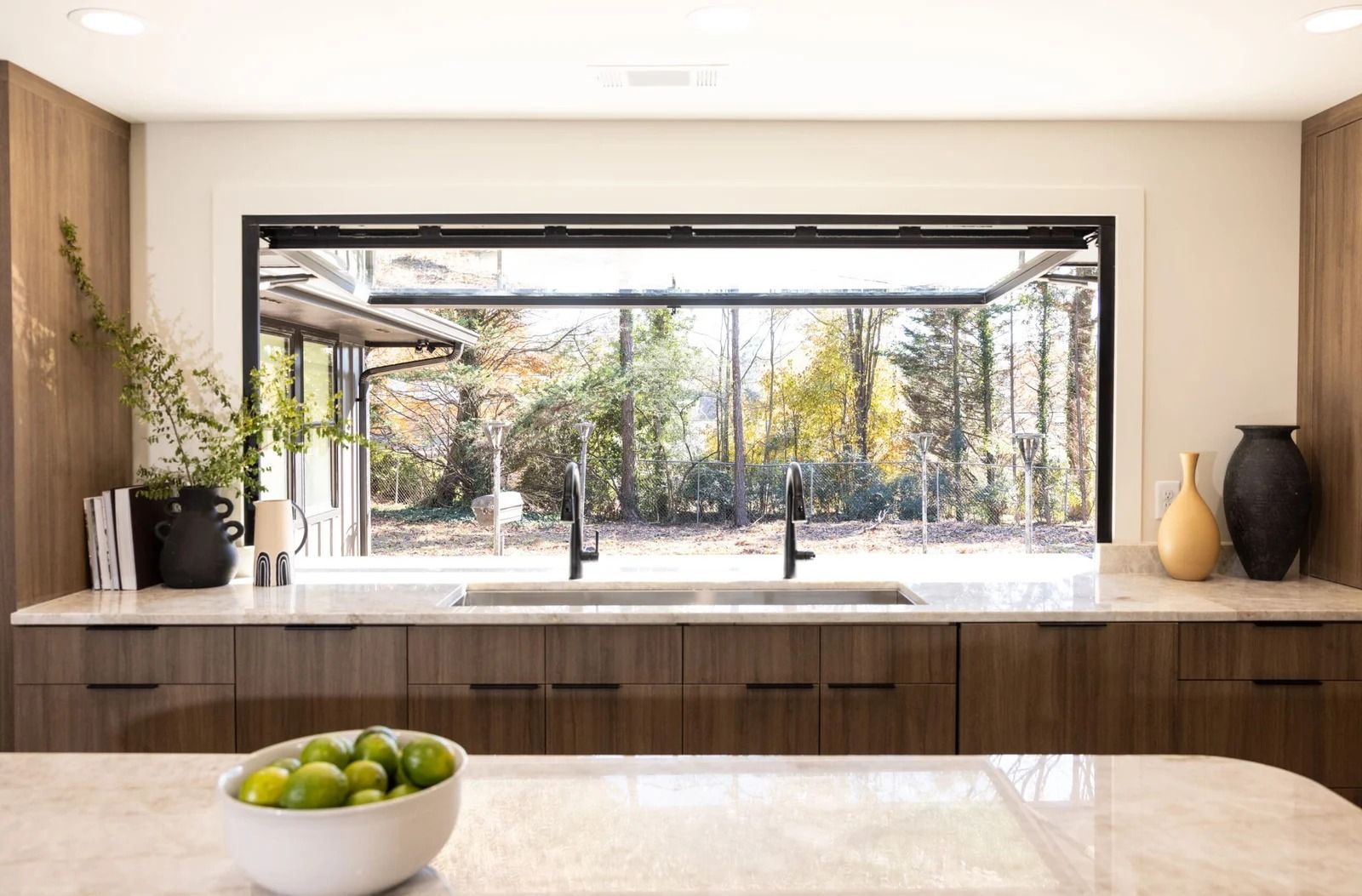 Modern kitchen with open window overlooking a wooded area. Brown cabinets, stone countertop, two black faucets.