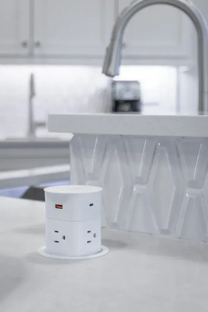White pop-up power outlet with USB ports on a light countertop, near a kitchen sink and faucet.