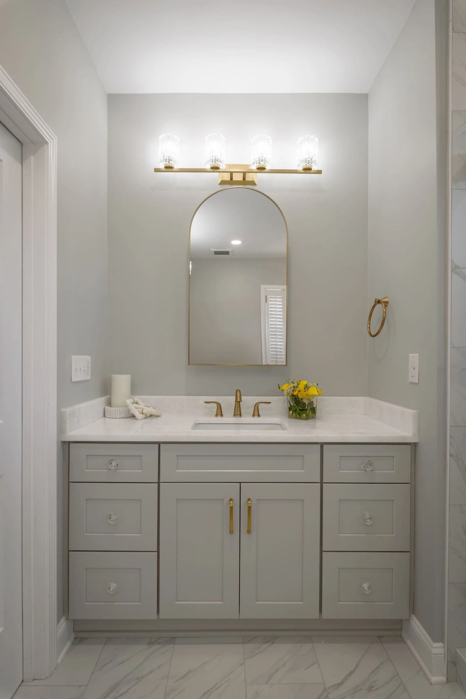 Gray bathroom vanity with gold accents, arched mirror, and white countertop.