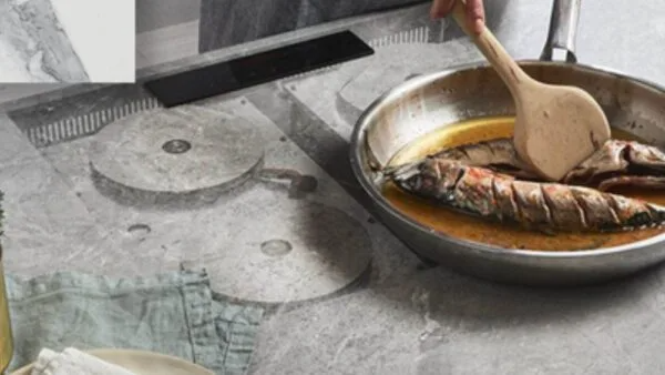 Fish cooking in a pan with a wooden spoon, on a textured countertop.