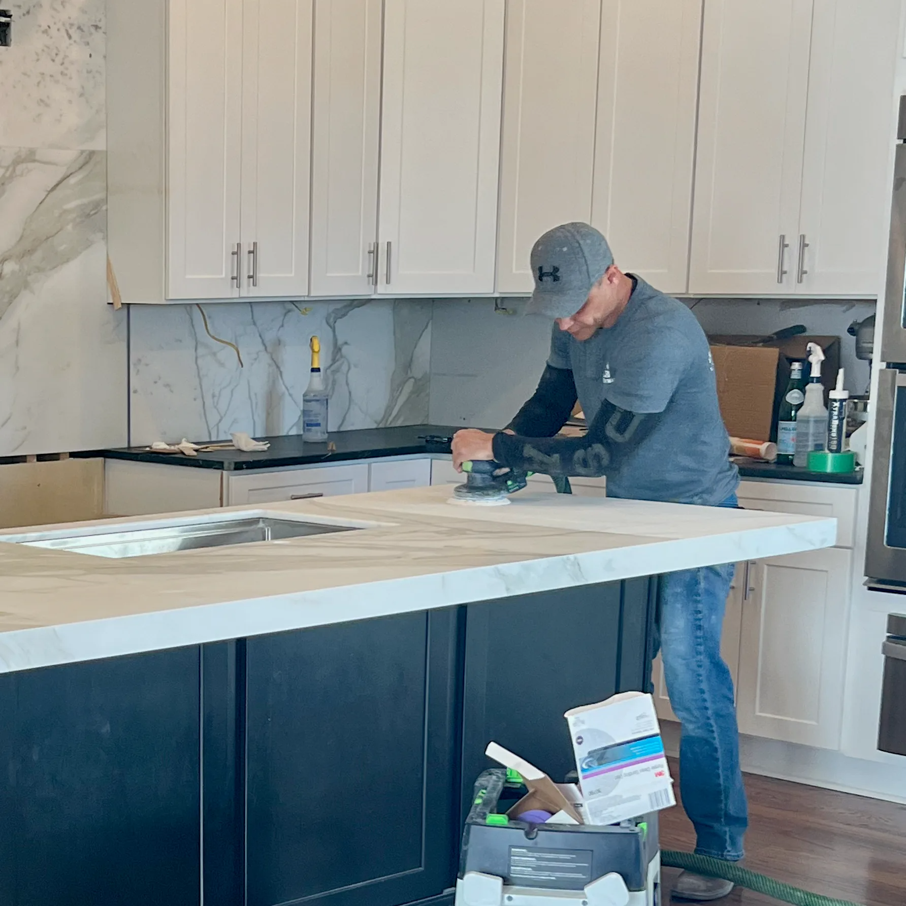 Man sanding countertop in a kitchen. Black cabinets, white uppers, marble backsplash. He wears a hat and jeans.