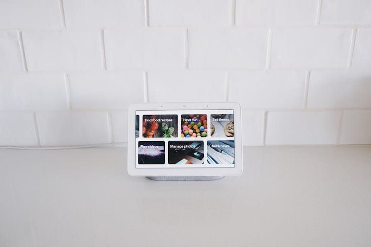 White smart display on a countertop against a white brick wall, showing food images on screen.