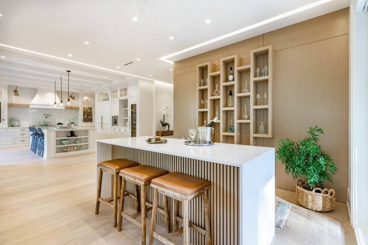 Modern kitchen with white island, three brown leather stools, and shelving unit with bottles.