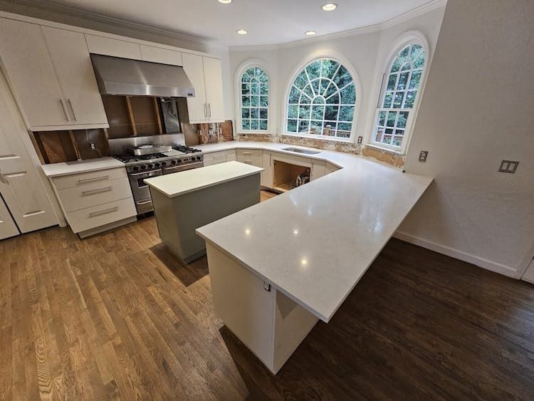 Kitchen with hardwood floors, white countertops, and arched windows overlooking trees.