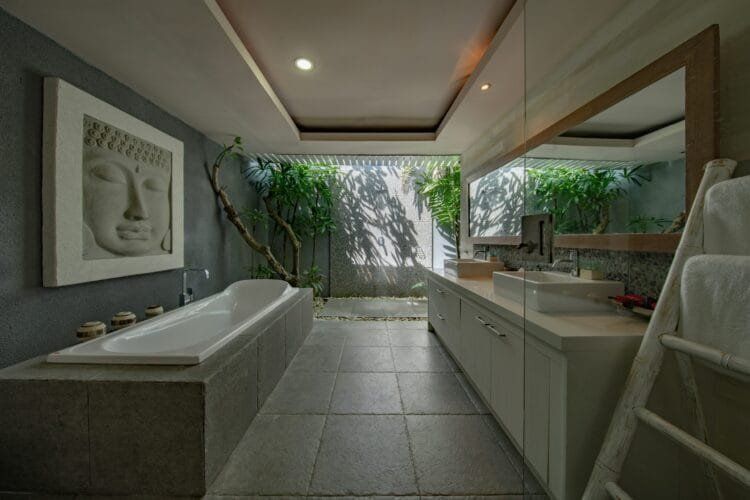 Spa-like bathroom with a bathtub, stone flooring, Buddha art, and natural light from greenery outside the window.