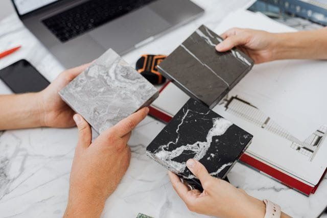 Three people holding up marble tile samples on a table. Laptop, phone, and blueprints visible.