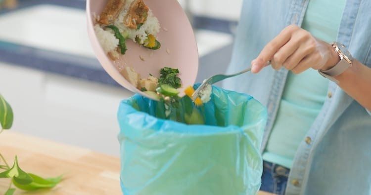 Person emptying food scraps from a pink pan into a blue trash bag in a kitchen.
