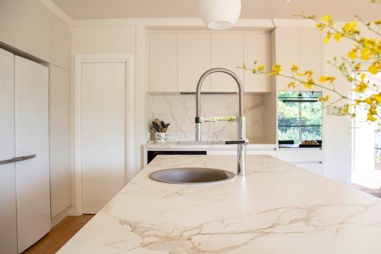 Modern white kitchen with marble countertops, stainless steel faucet, and yellow flower arrangement.