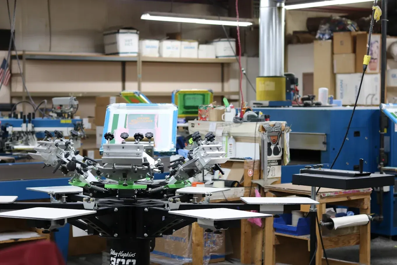 Screen printing machine in a workshop, surrounded by equipment and boxes.