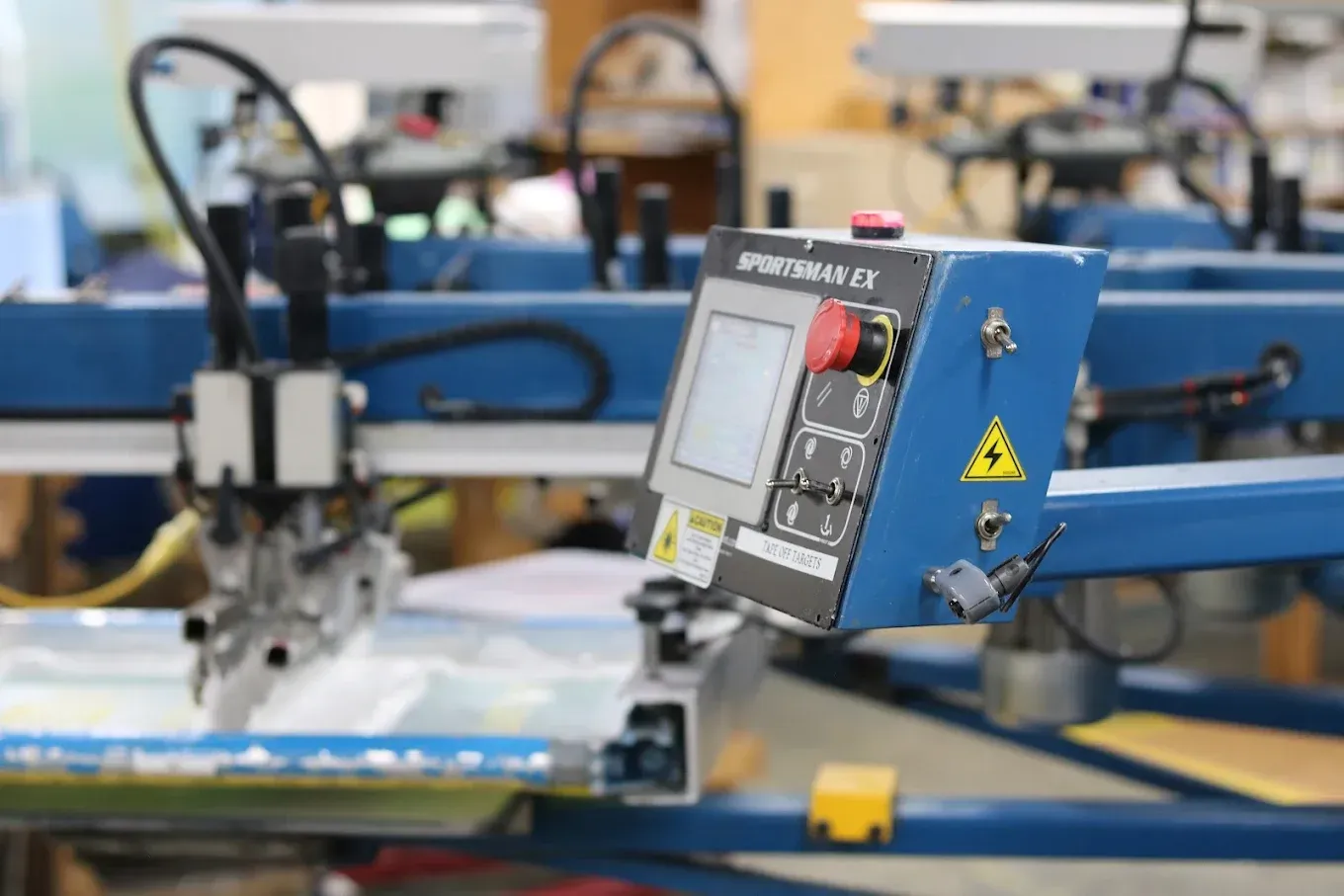 Control panel on blue screen printing machine. Large red stop button and small screen visible.