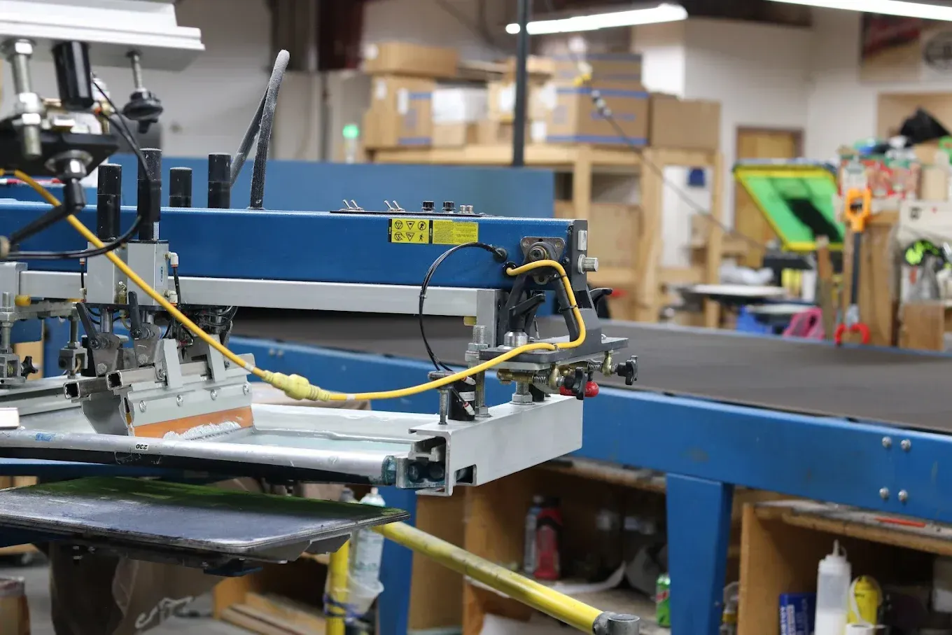 Screen printing machine in a workshop, blue and yellow components, boxes in the background.