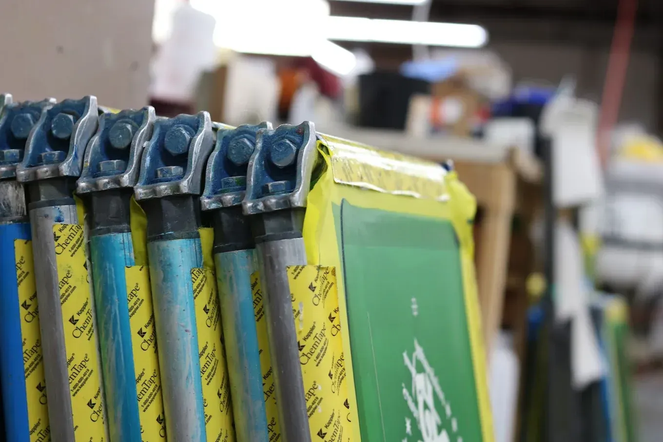 Row of screen printing frames with blue and yellow handles, in a workshop setting.