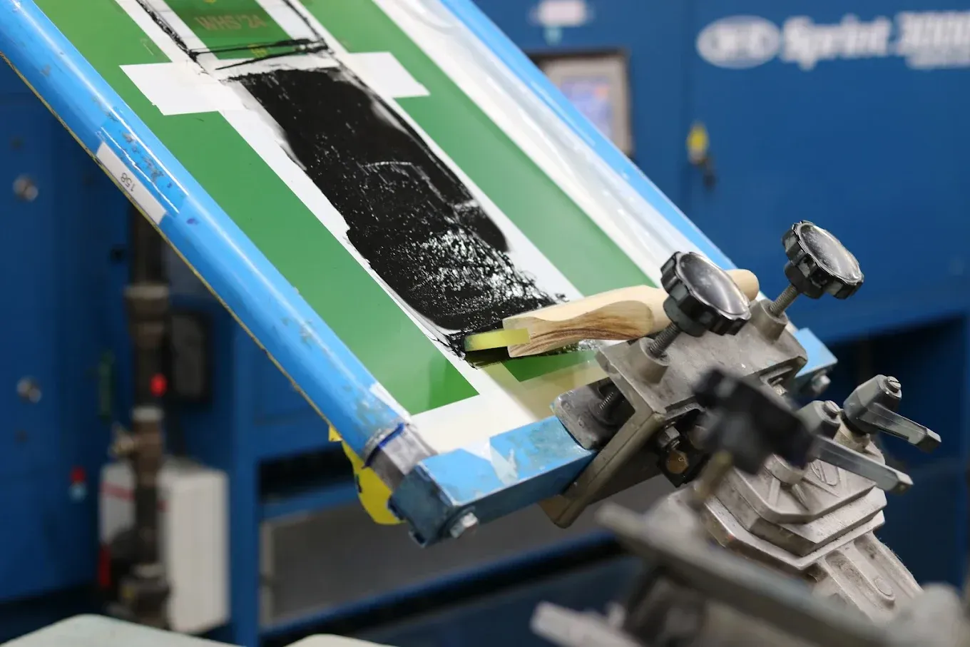 Screen printing machine with a stencil, applying black ink onto a white and green surface.
