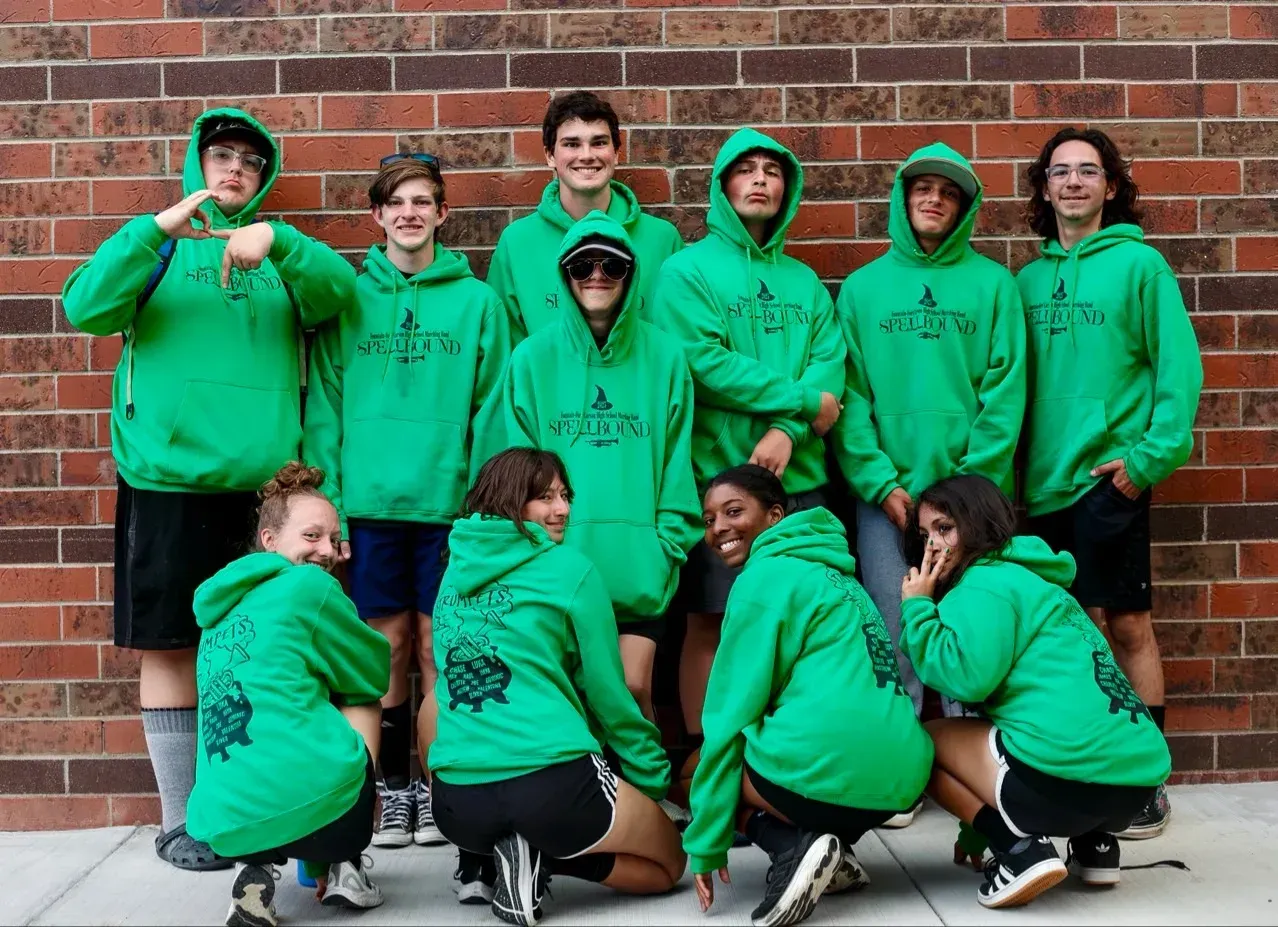 Group of people in green hoodies pose in front of a brick wall; some kneel, some point, all wear matching designs.