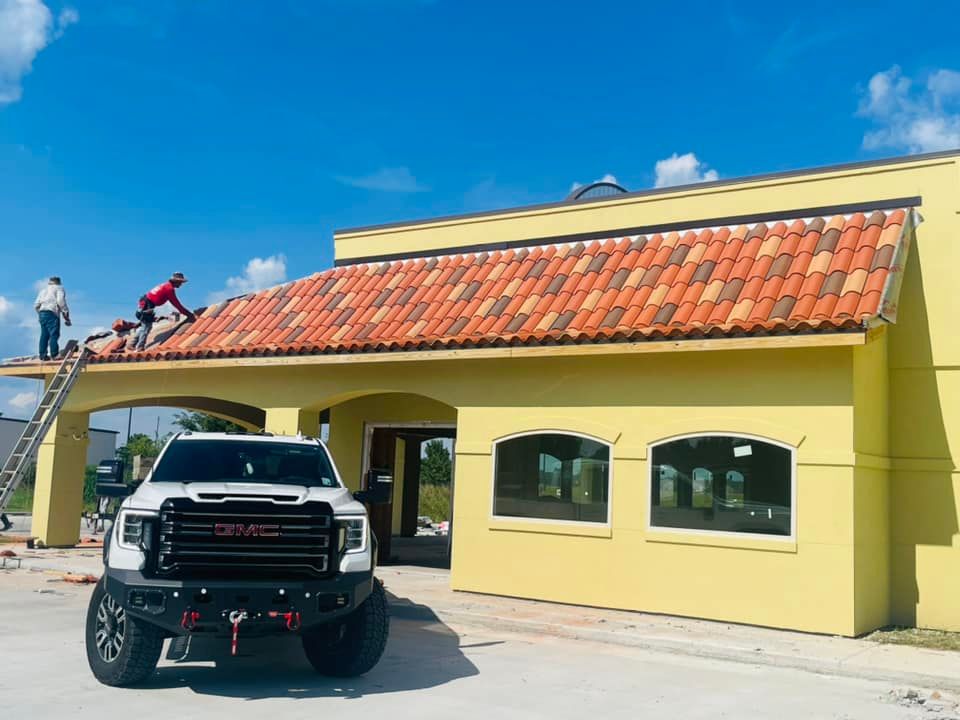 Yellow house with terracotta roof, lifted black truck in front, and workers on the roof under a blue sky